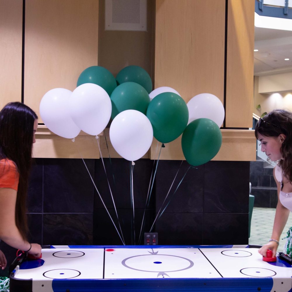 Two students play air hockey in front of green and white balloons in the Student Union near Charlotte Barnes & Noble