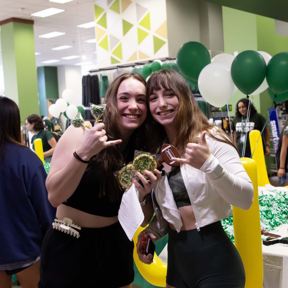 Two students pose with picks-up hand signs and hold cookies with green and white sprinkles in Charlotte Barnes & Noble