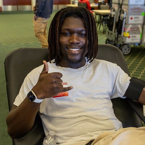 Smiling student gives blood and puts his hand up like a pick axe