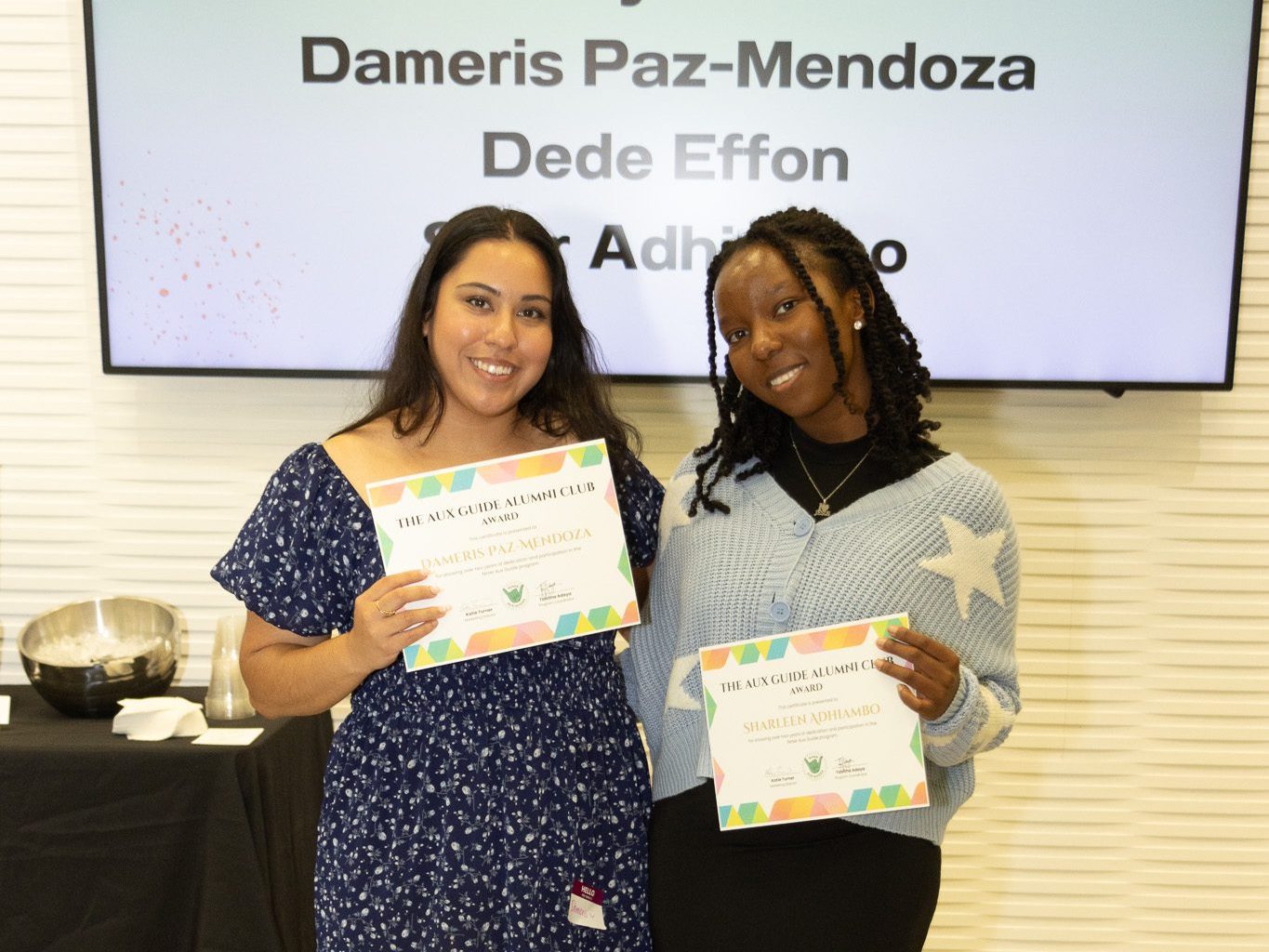Dameris Paz Mendoza and Sharleen Adhiambo smile and hold their Aux Guide Alumni Club certificates.