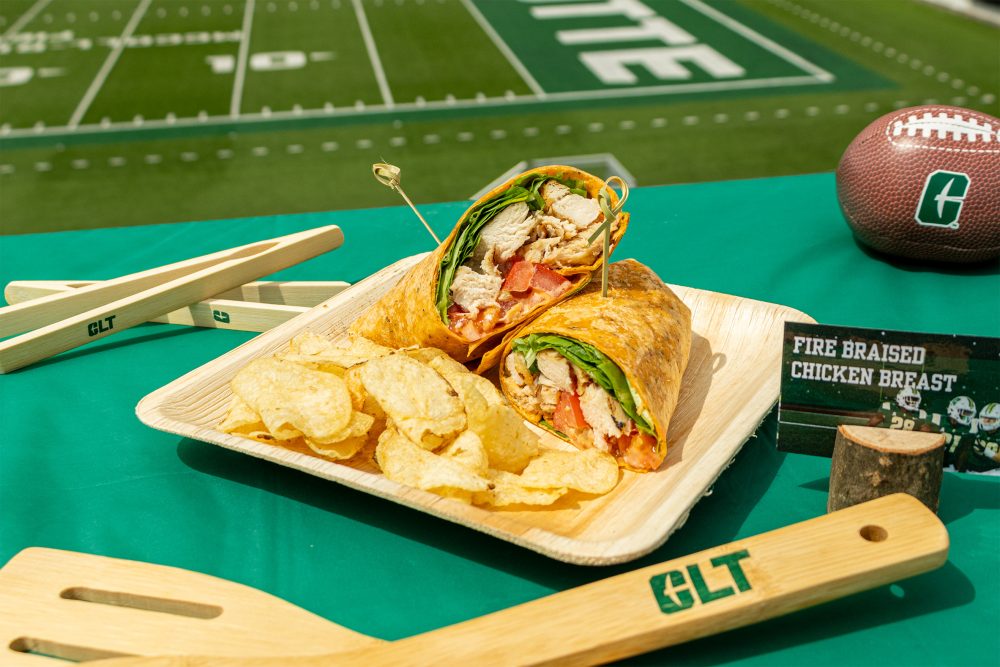 sandwich wrap with chicken, spinach and tomatoes sits on a plate with potato chips, posed along with side that reads "fire braised chicken breast" with Charlotte's Jerry Richardson stadium football field in the background