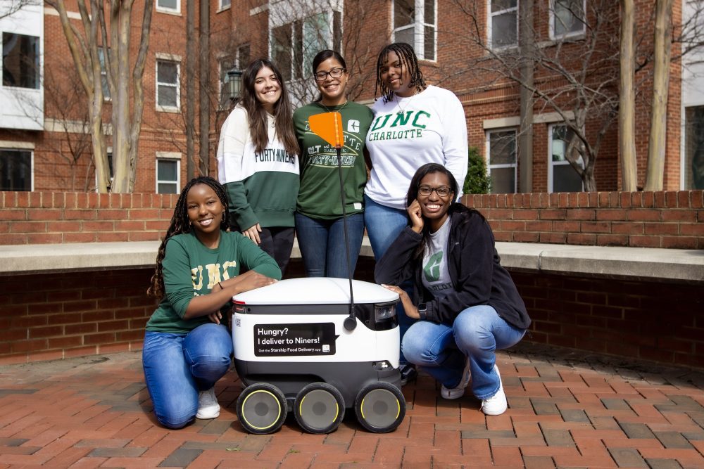Smiling students wearing Charlotte t-shirts and sweaters pose with a Starship delivery robot