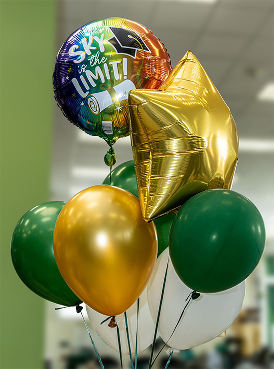 balloon bouquet with a gold star-shaped balloon, rainbow balloon that reads "the sky is the limit!" and several green and gold latex balloons