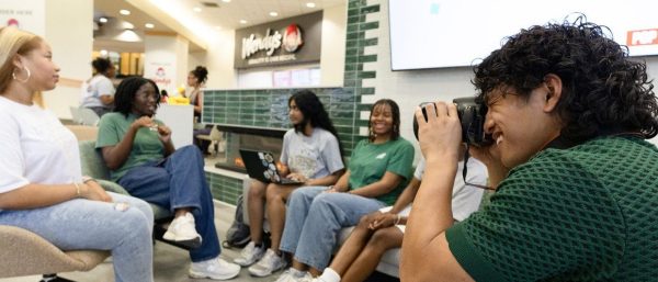 smiling student intern wearing a green polo holds a camera to his eye, taking a picture of a group of students sitting in a circle in the Student Union food court